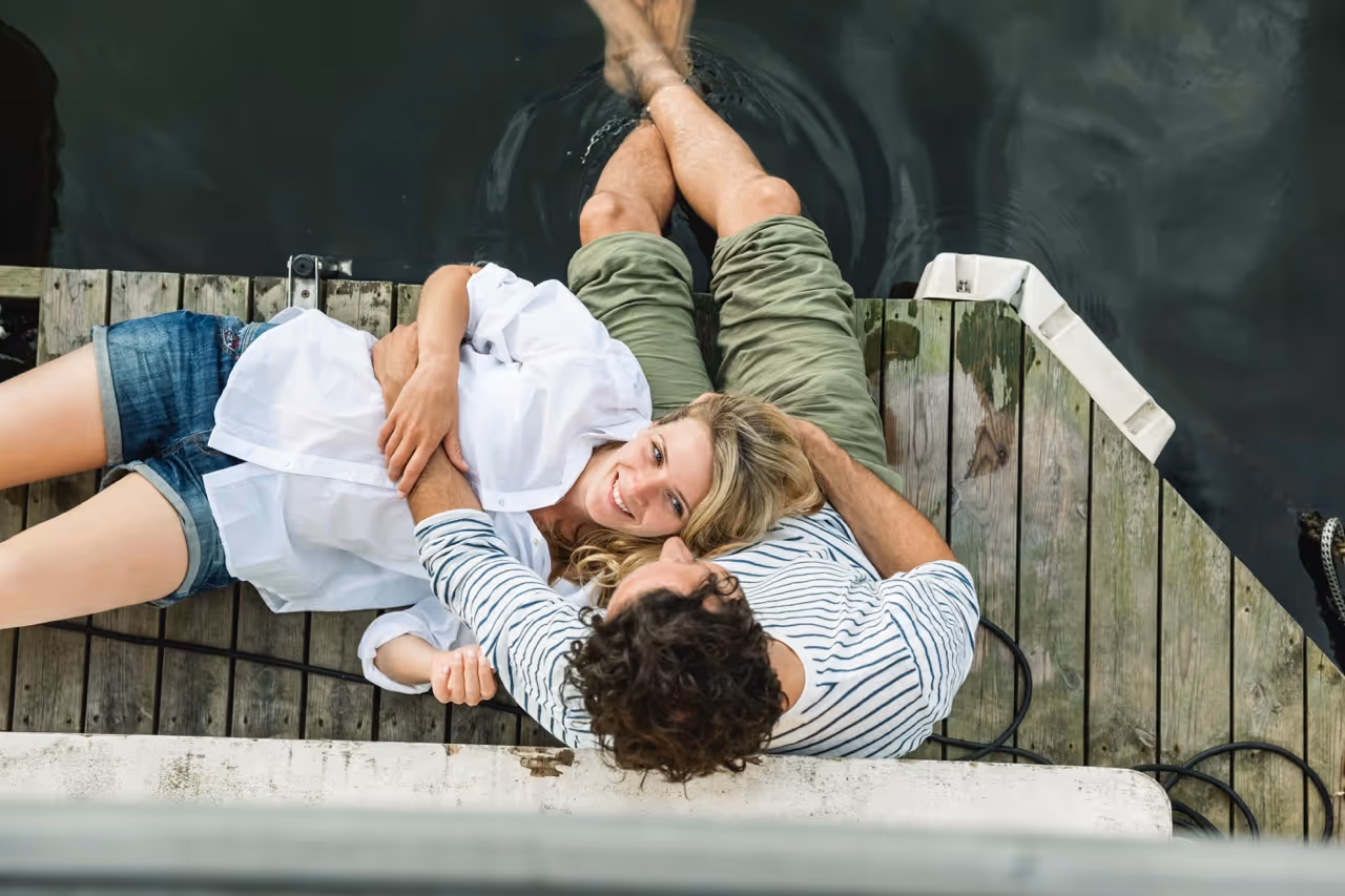 Couple relaxing on a wooden dock with the woman lying on the man’s lap near calm water.