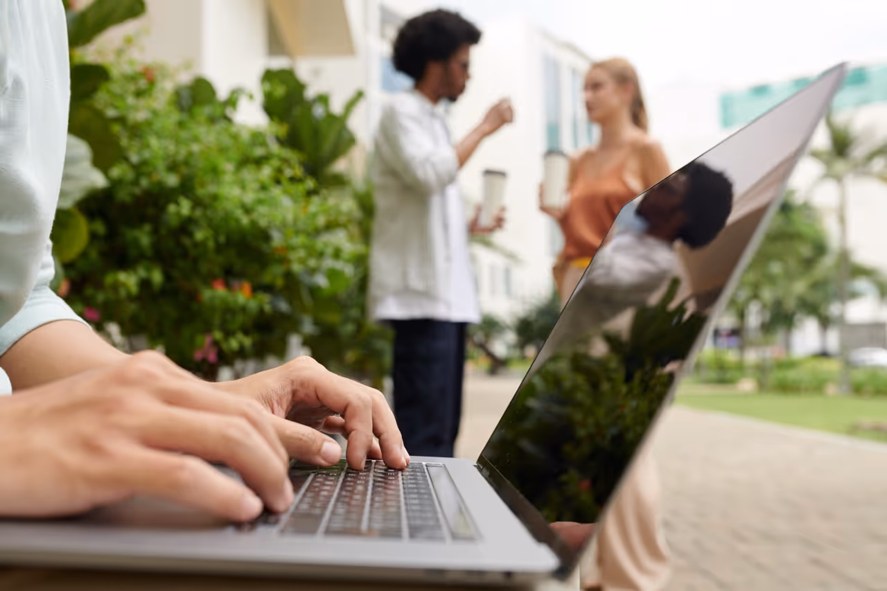 Close-up of hands typing on a laptop outdoors with two people blurred in the background holding coffee cups.