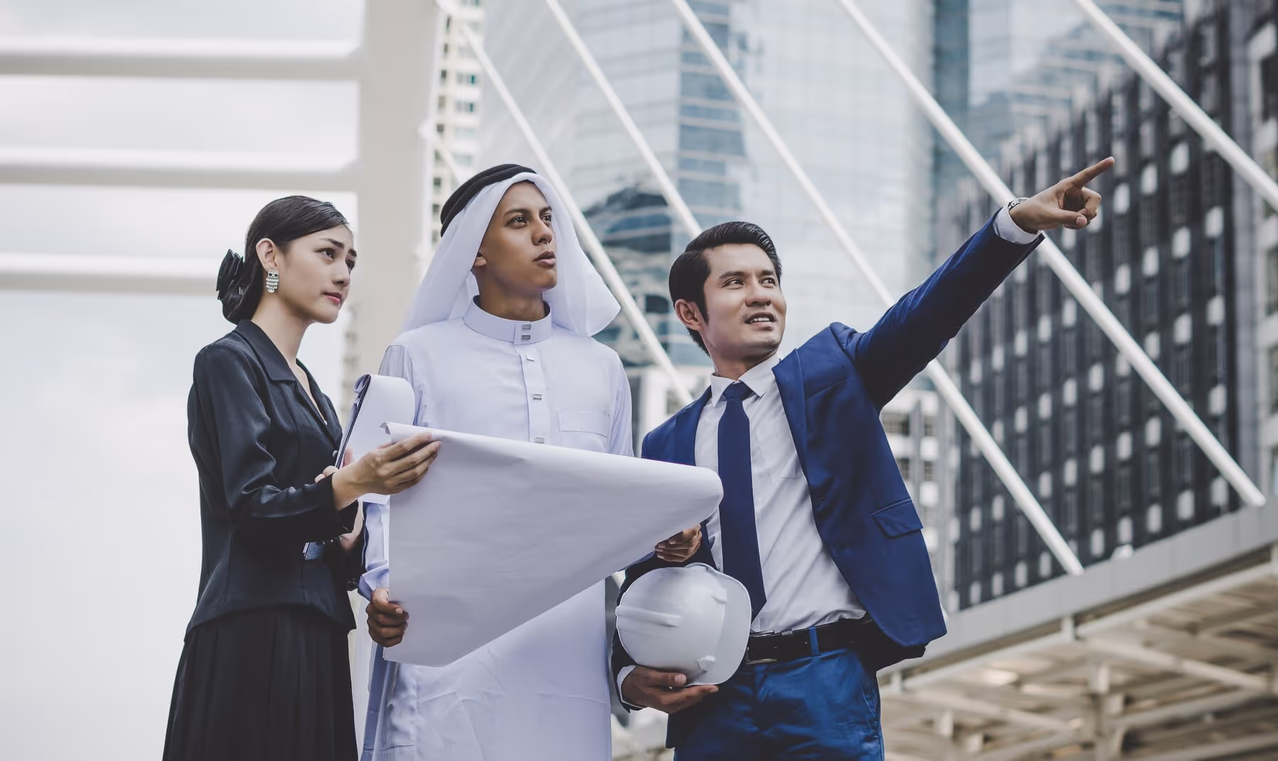 Three real estate professionals, one in traditional Emirati attire and two in business suits, stand outdoors with blueprints, with one pointing towards a modern building, illustrating the collaborative effort to sell property in Dubai.
