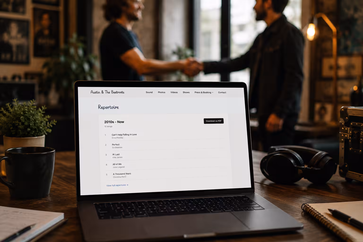 Laptop on wooden table shows music repertoire list while two men shake hands in blurred background.
