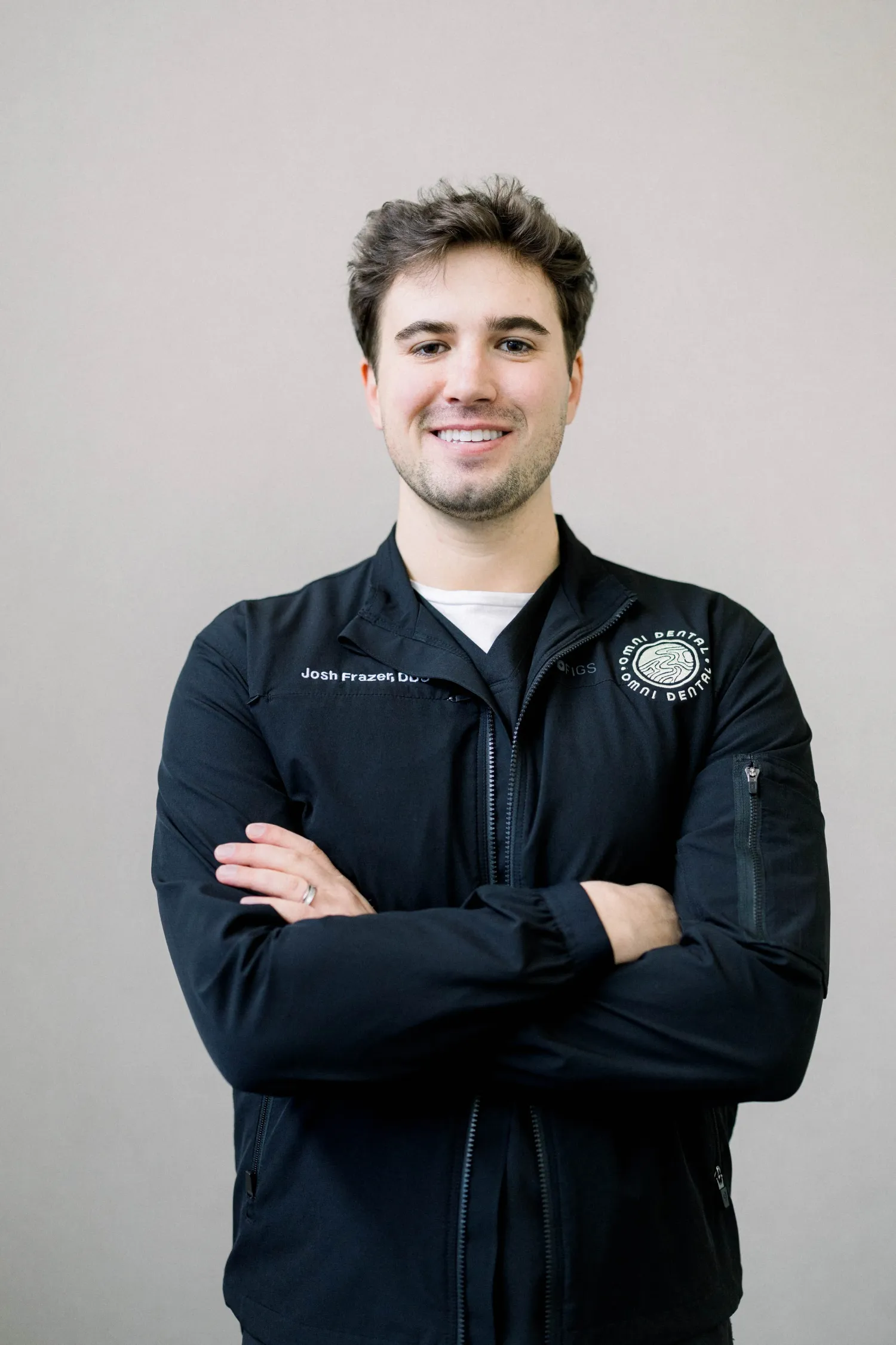 Man in black jacket with Omni Dental logo smiling with arms crossed against a plain background.