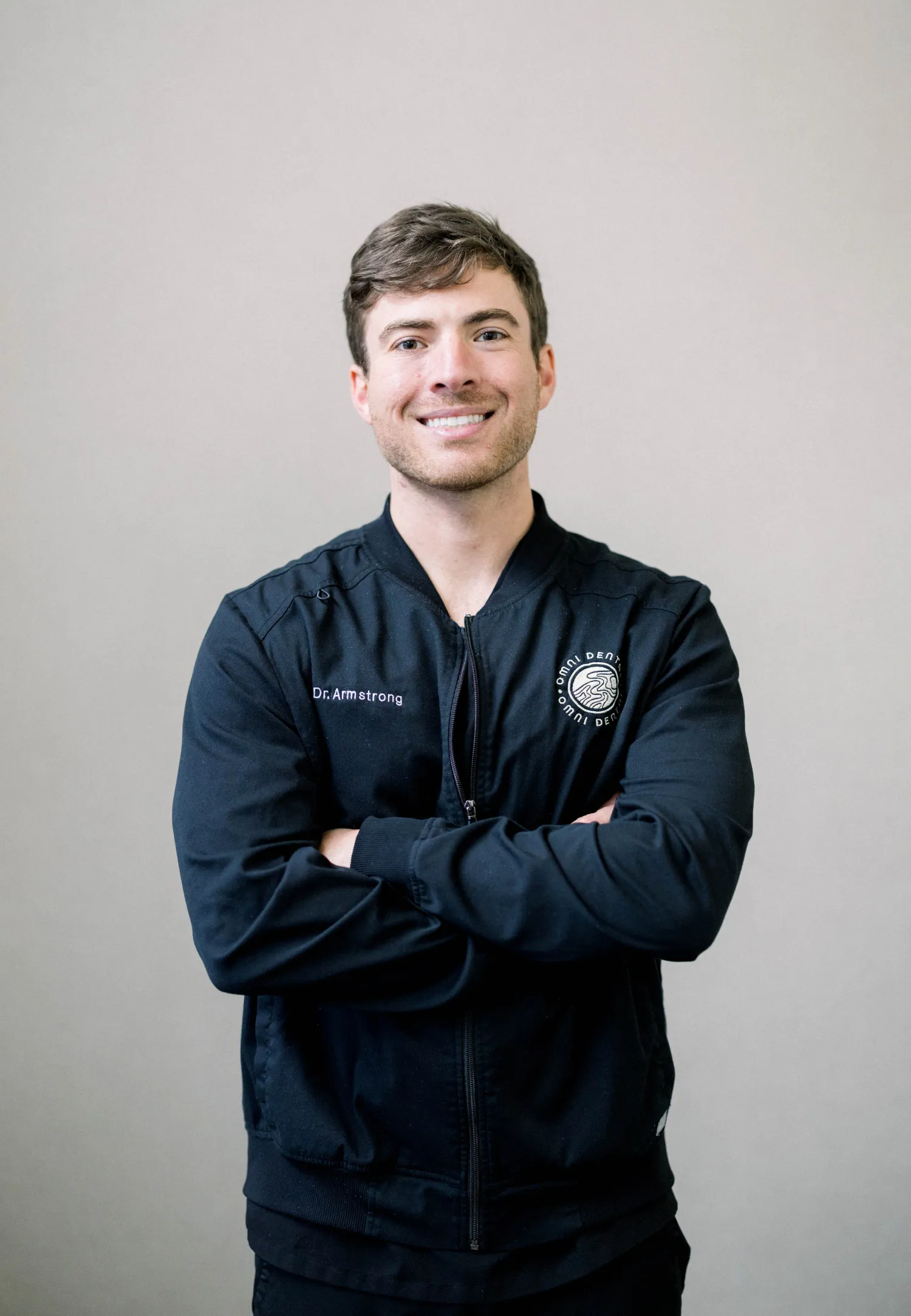 Man with short brown hair smiling, wearing black jacket reading 'Dr. Armstrong' and standing with arms crossed.