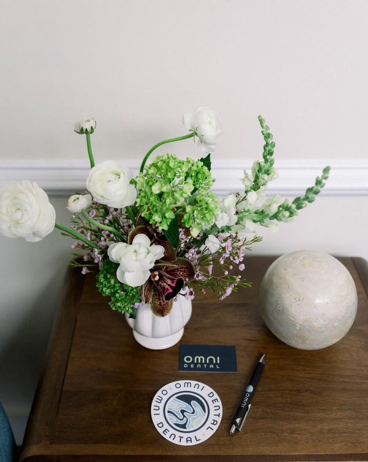 Floral arrangement in white vase on wood table with Omni Dental card, coaster, and pen beside a stone sphere.