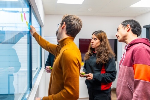 Drei Personen kleben bunte Notizzettel an eine Glaswand in einem Büro.