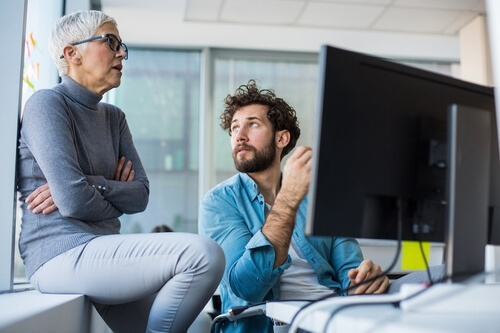 Ältere Frau und jüngerer Mann diskutieren vor einem Computerbildschirm in einem modernen Büro.