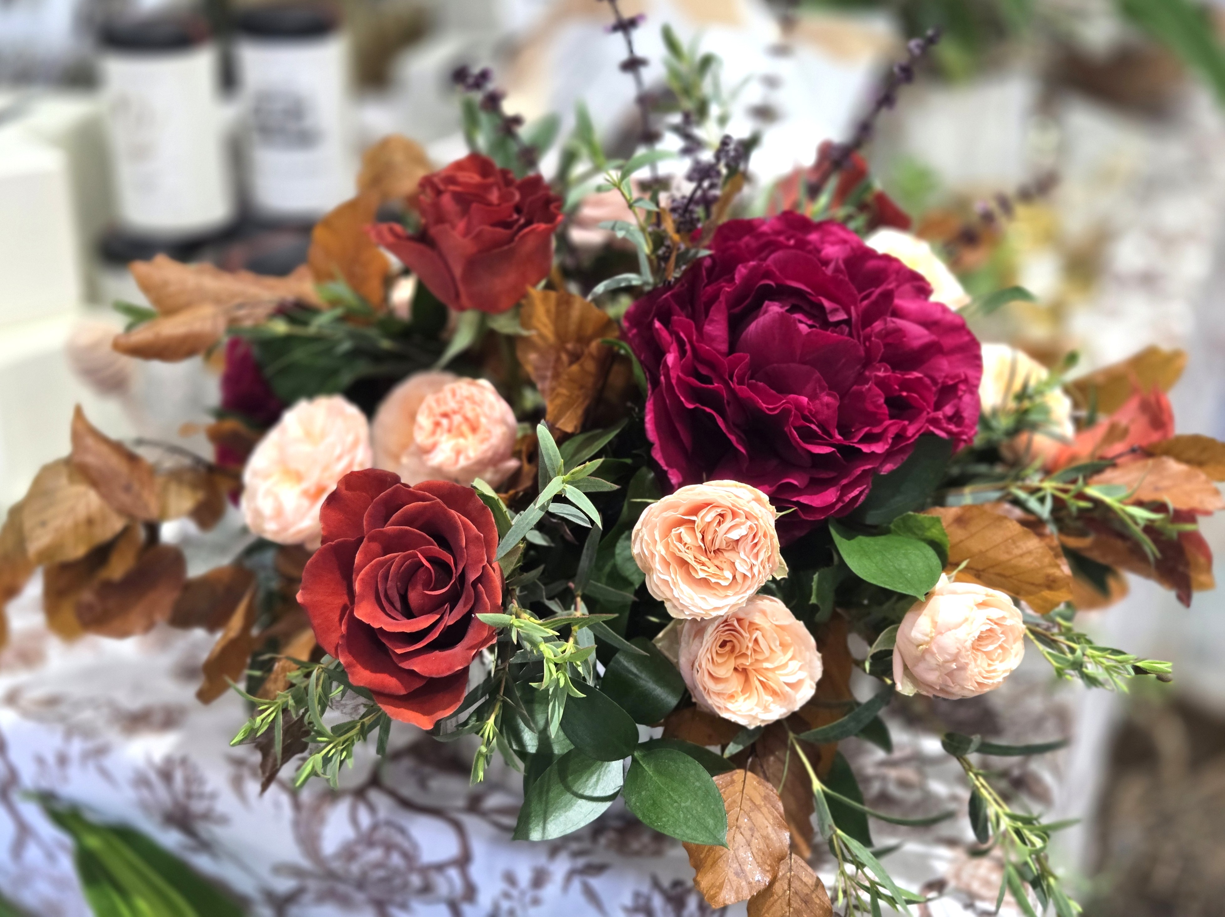 Thanksgiving floral centerpiece featuring peach garden roses, burgundy blooms, burnt orange roses, autumn foliage, and lush greenery arranged in an organic, elegant style.