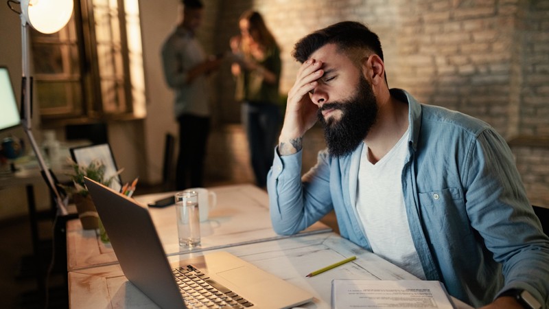 A business owner sitting at a desk and holding his head in stress while looking at his laptop, symbolizing fixed price project overwhelm