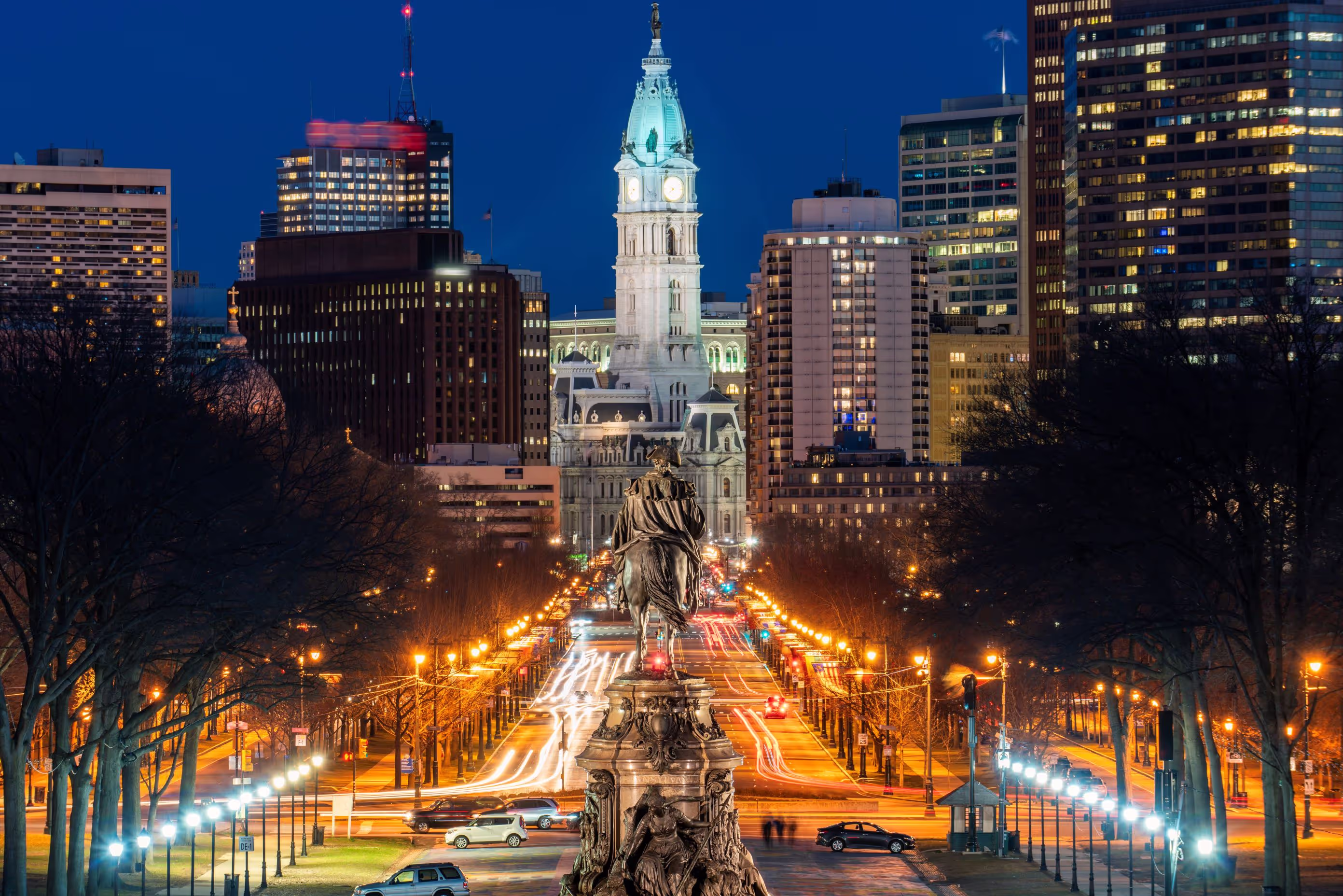 Night view of Philadelphia cityscape with illuminated City Hall and a statue in the foreground along a busy street with light trails.