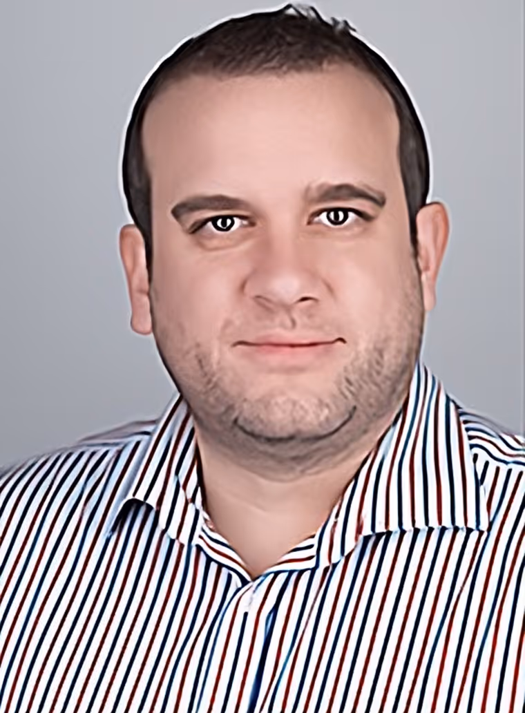 Man with short dark hair wearing a white shirt with vertical red and blue stripes against a plain background.