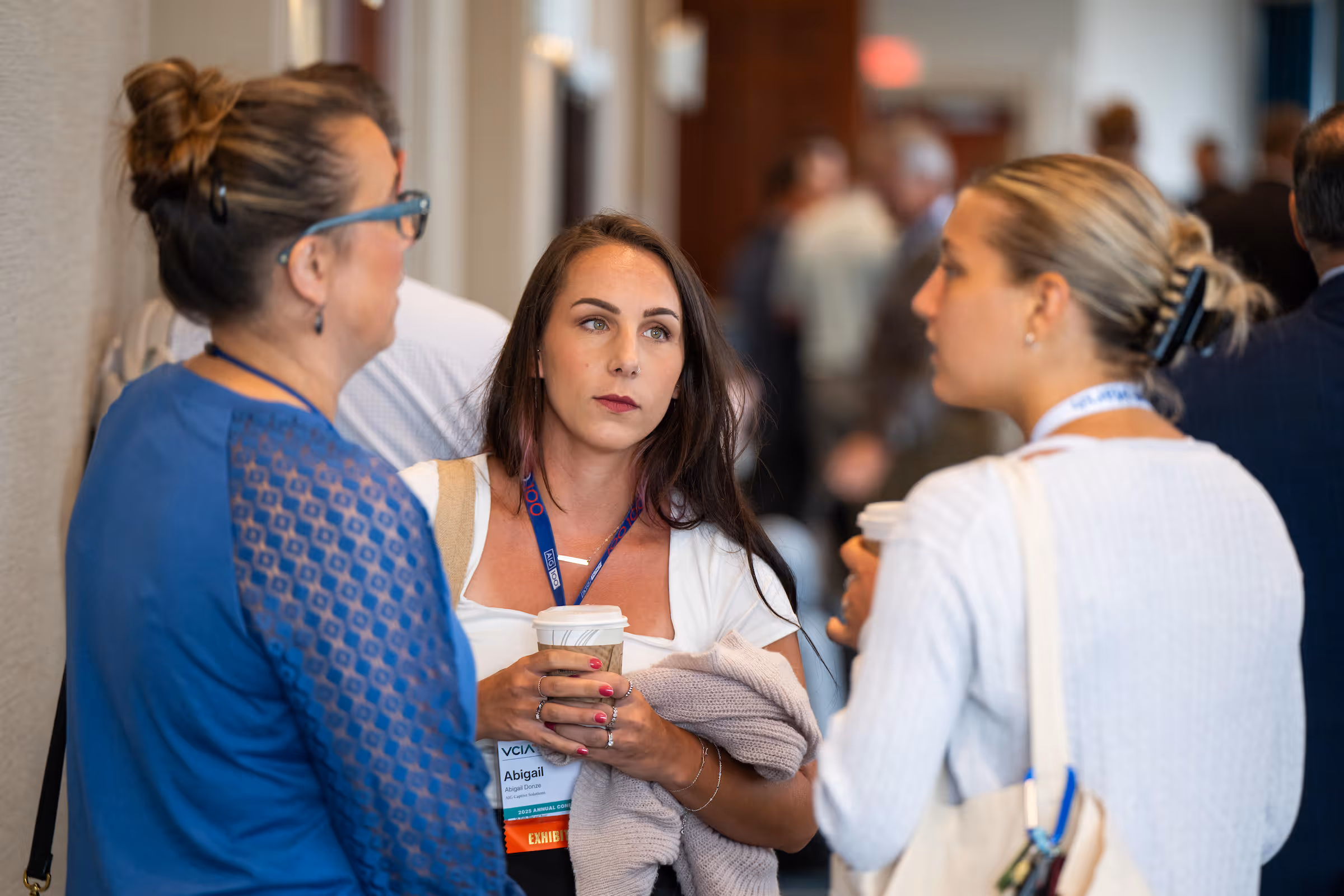 Three women engaged in conversation at a captive insurance conference, holding coffee cups and wearing name badges.