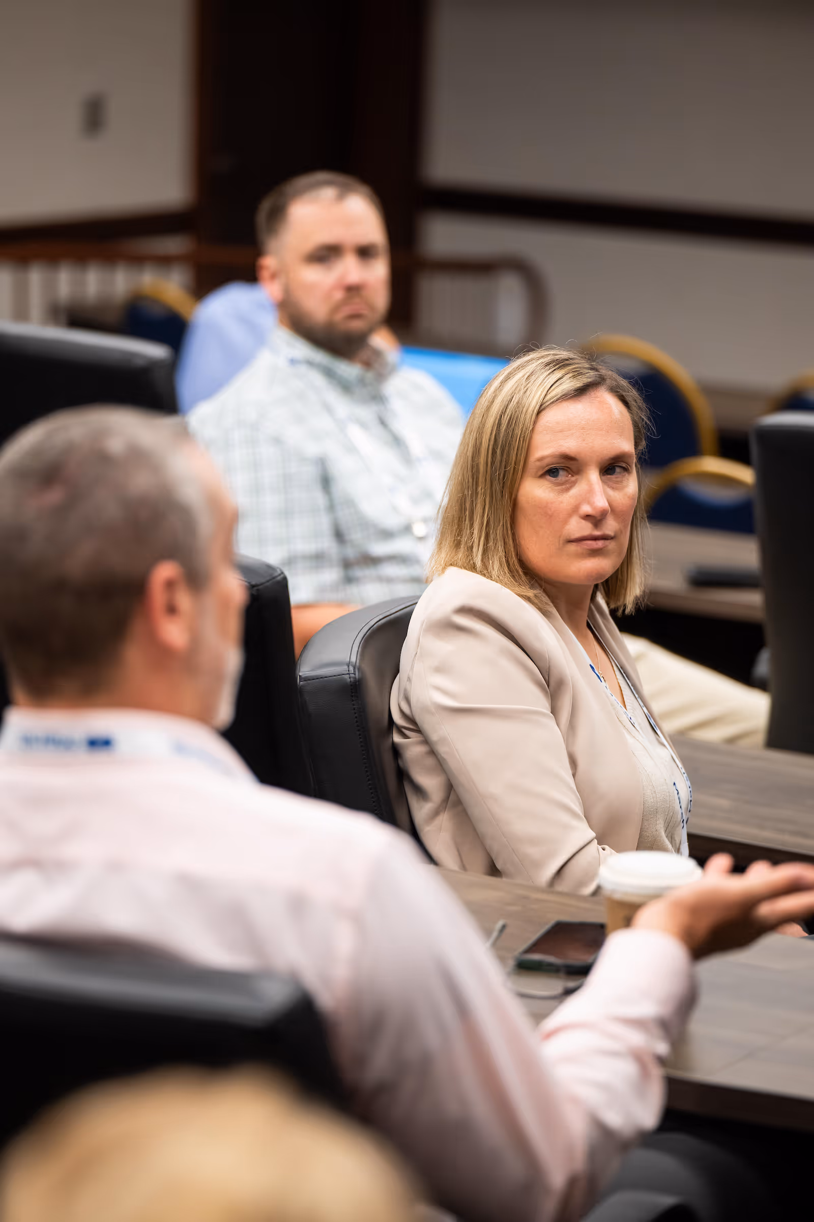 Woman in a beige blazer attentively listening during a meeting with two men in a conference room.