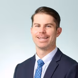 Smiling man with short brown hair wearing a navy suit, white shirt, and blue patterned tie against a light gray background.