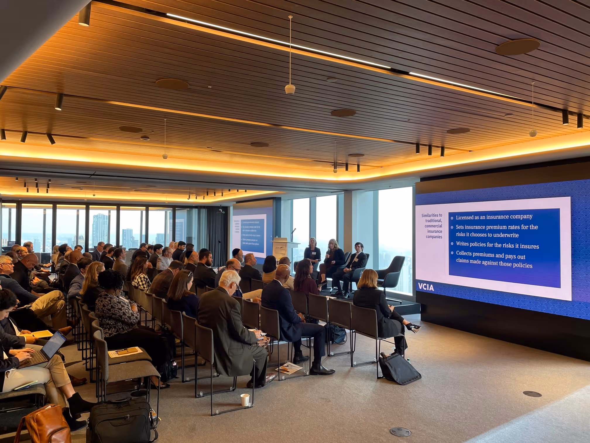 Audience seated watching a panel discussion in a modern conference room with large windows and a presentation slide on captive insurance company similarities displayed on a screen.