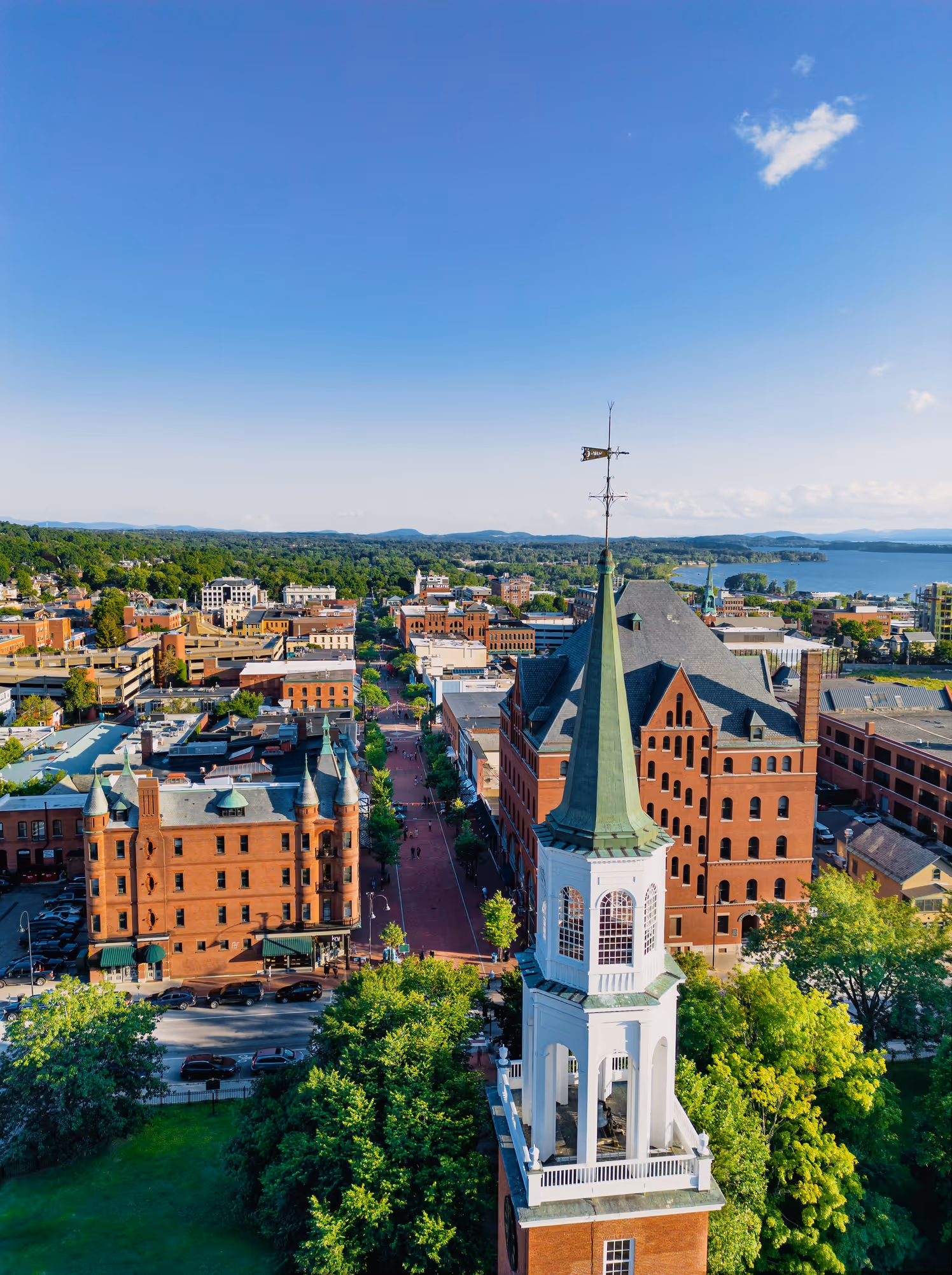 Aerial view of Burlington, Vermont, with a tall church steeple in the foreground, red brick buildings, tree-lined streets, and Lake Champlain in the background under a clear blue sky.
