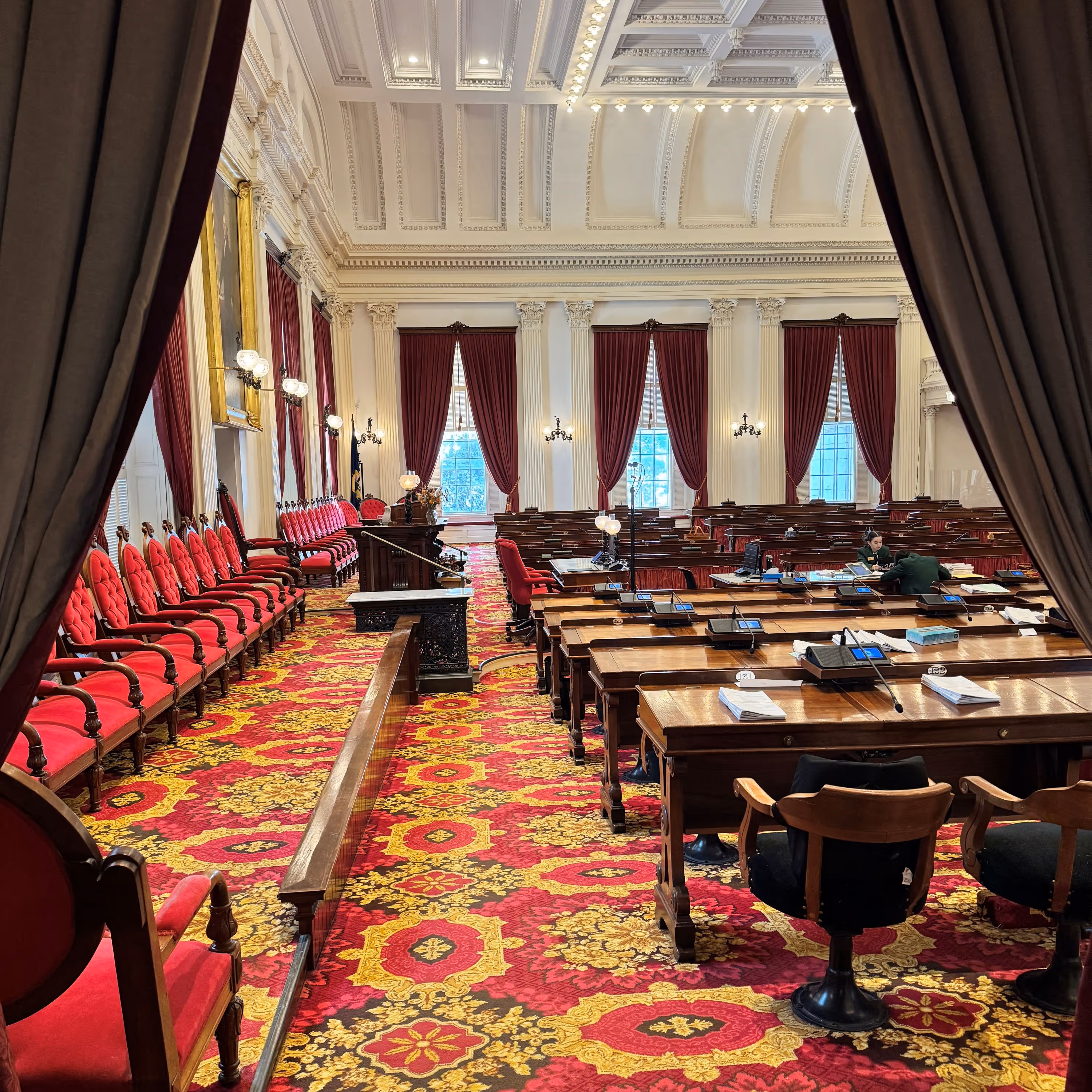 View into an ornate legislative chamber with red velvet chairs, wooden desks, and large windows framed by red curtains.