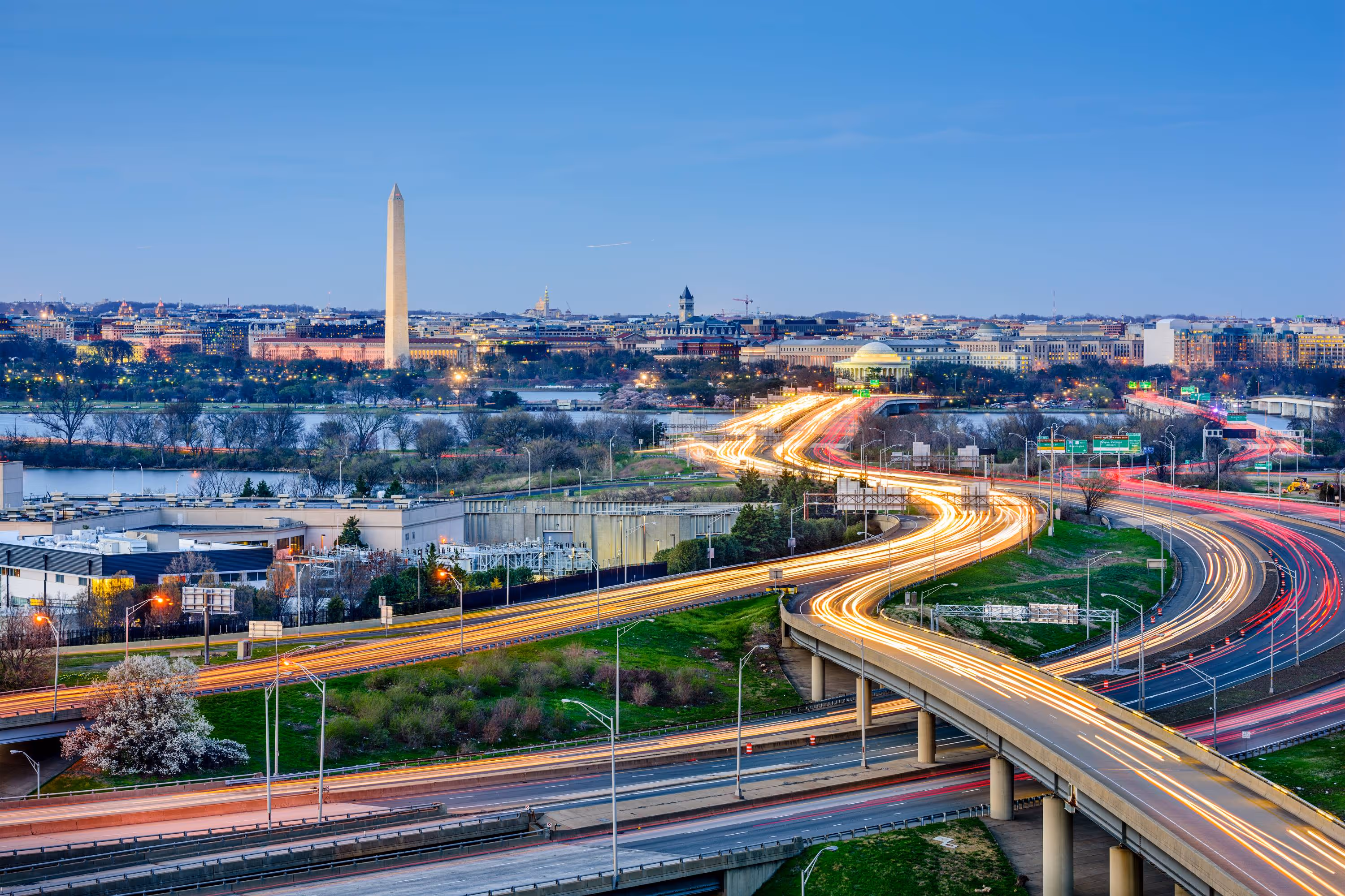 Long-exposure view of highways with light trails leading to the Washington Monument and Jefferson Memorial in Washington, D.C. at dusk.