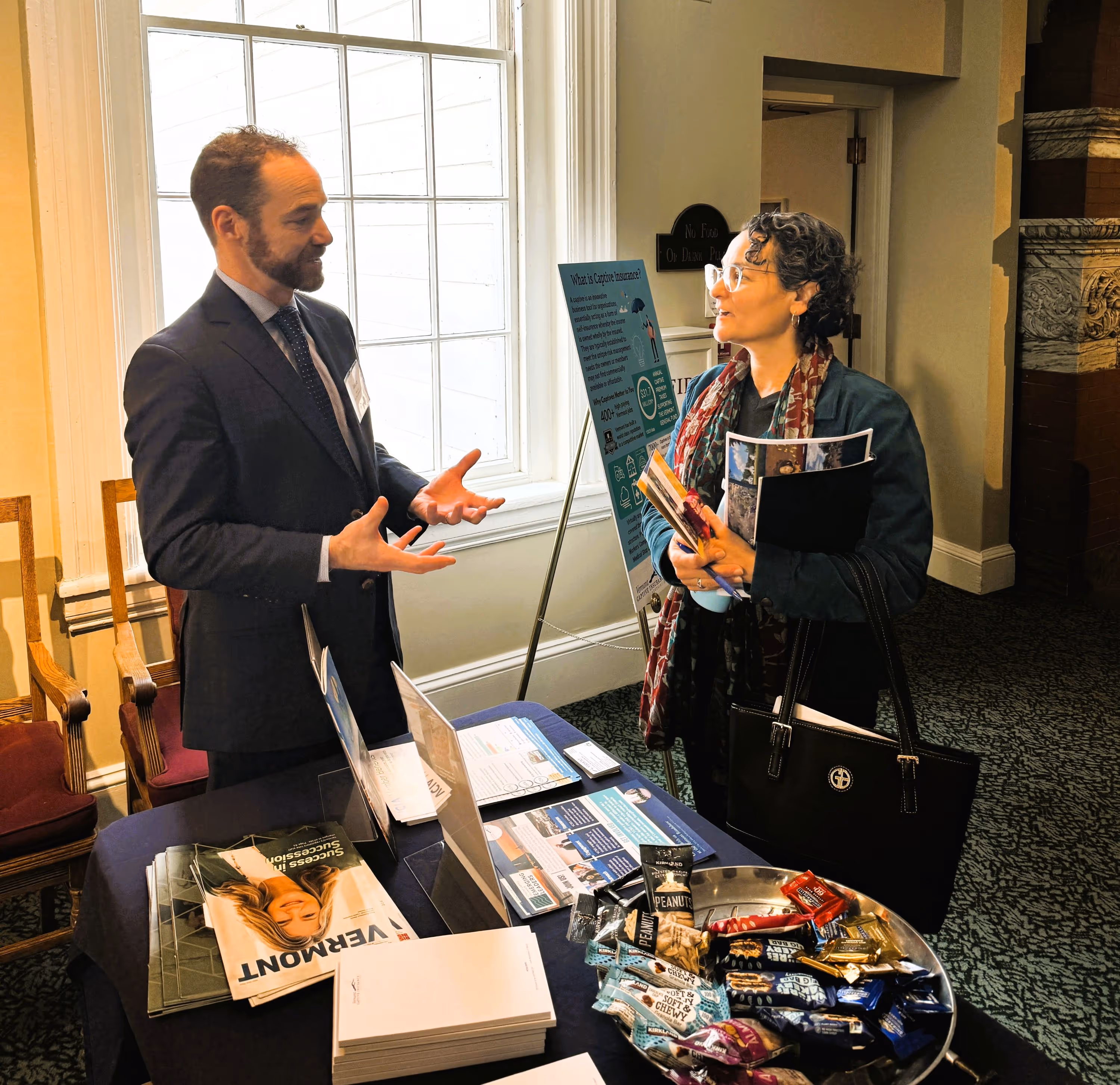 Captive insurance professional in suit explaining information at a table to a lawmaker holding brochures and a bag in a well-lit indoor setting.