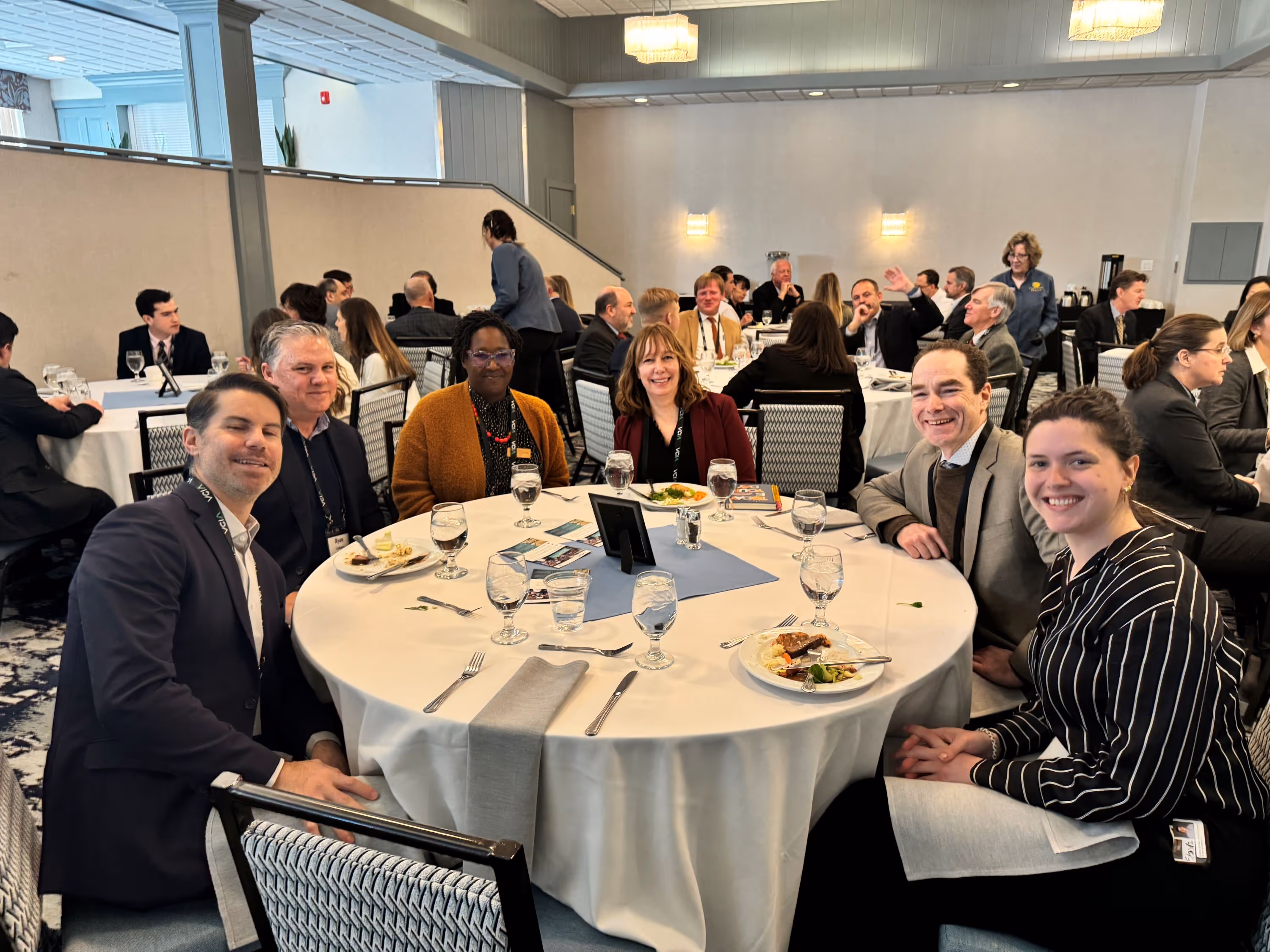 Six people sitting around a round table in a banquet hall, smiling with plates of food and water glasses in front of them.