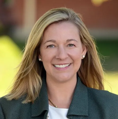 Smiling blonde woman with light eyes wearing a dark green jacket and white top, outdoors with blurred background.