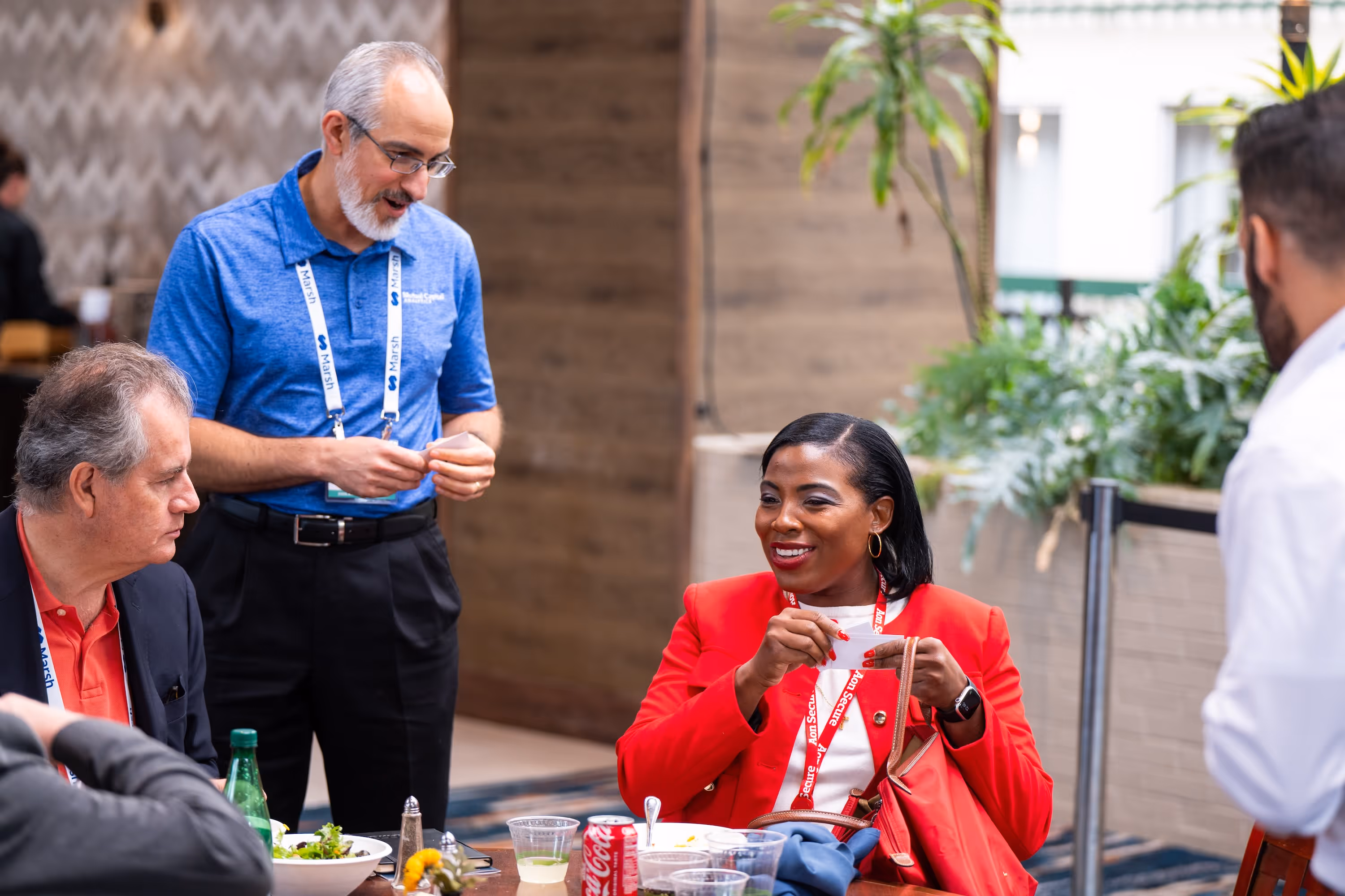 A woman at the VCIA Conference in a red jacket smiling and holding a paper slip while talking with three men around a table with food and drinks.