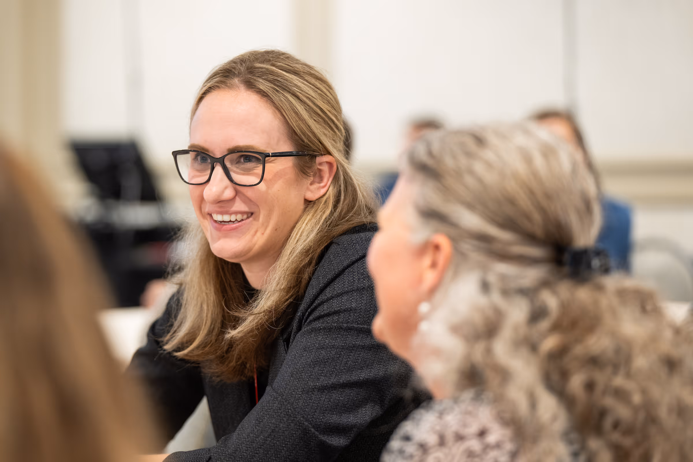 Smiling woman with glasses and long blonde hair engaged in conversation with another person in a meeting setting.