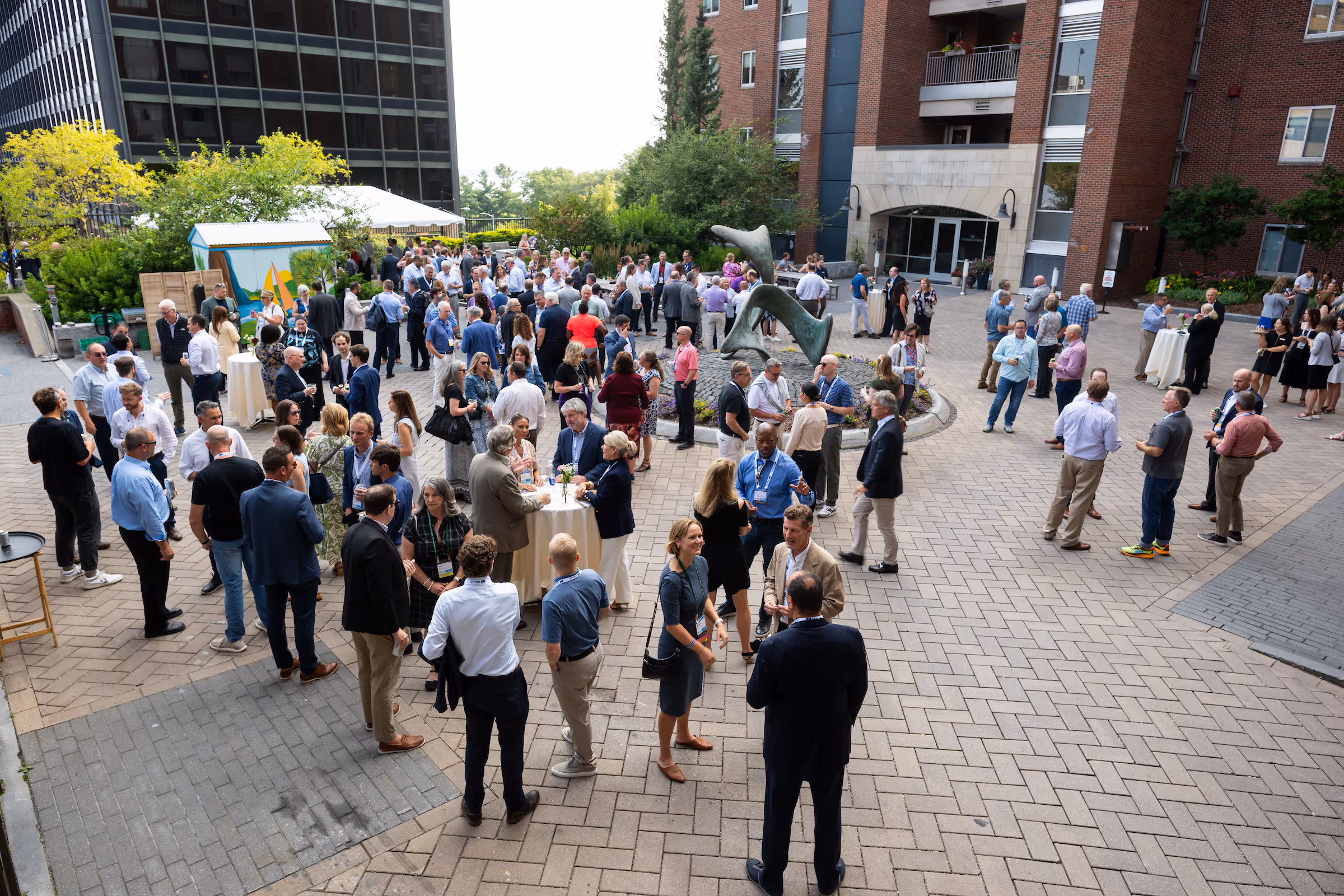 Large group of people socializing outdoors at a formal event near a modern building with a sculpture in the center.