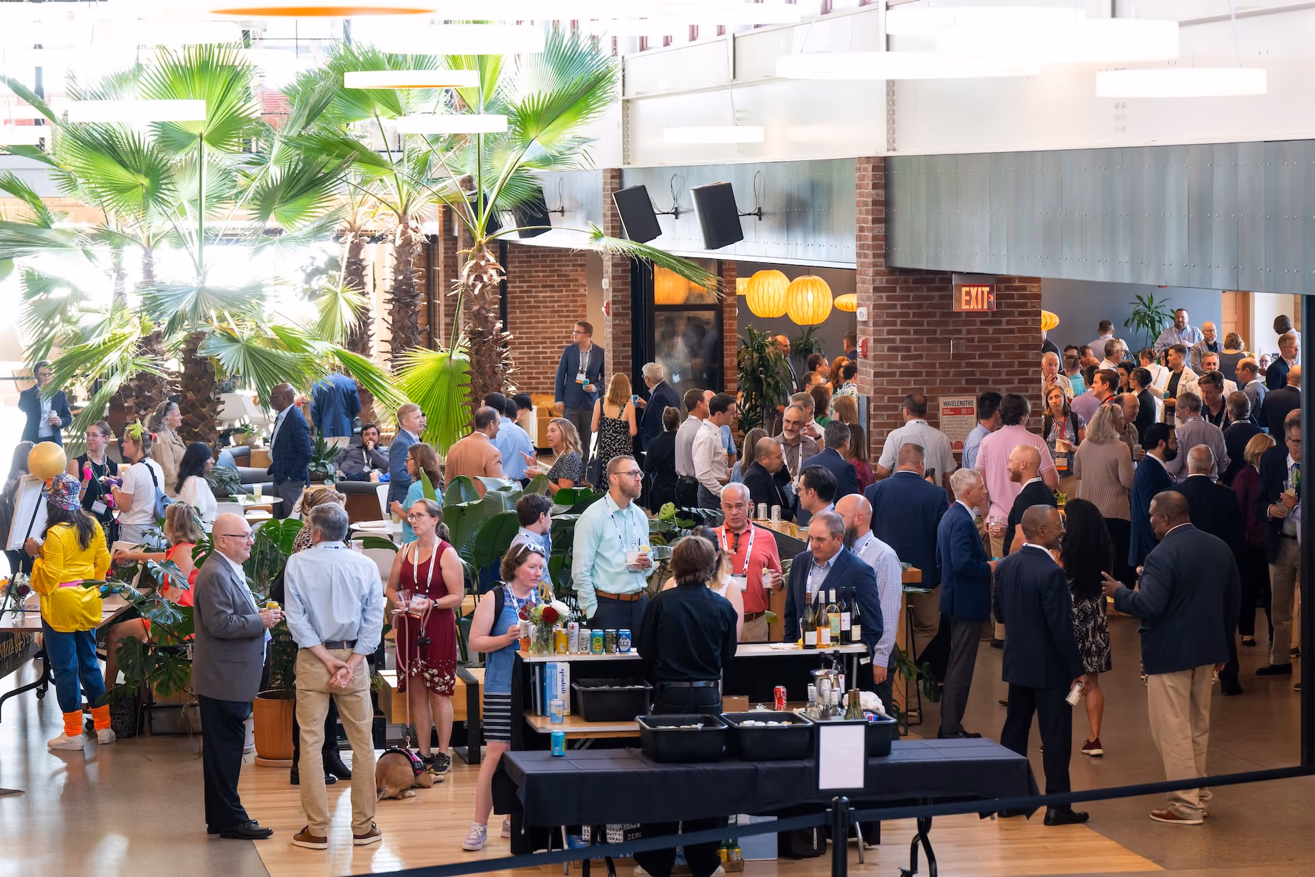 Large indoor networking event with diverse attendees socializing near a bar area surrounded by tropical plants and modern decor.