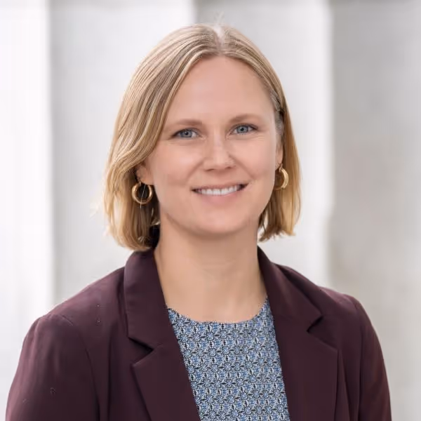 Smiling woman with short blonde hair wearing gold hoop earrings, a patterned blue top, and a dark blazer.