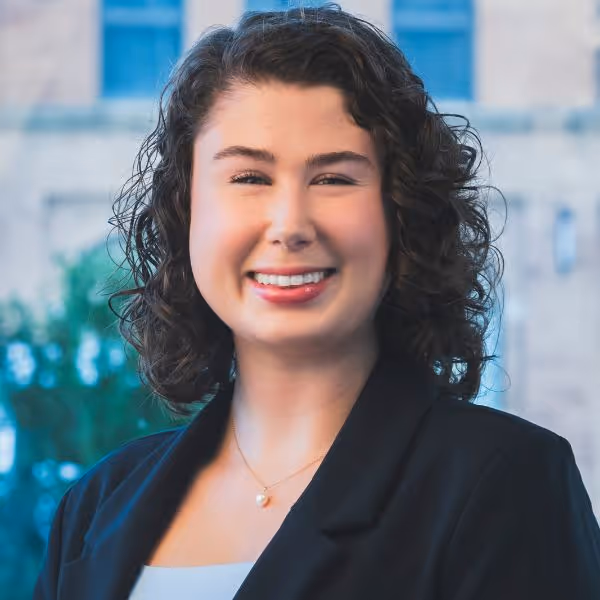 Smiling woman with curly dark hair wearing a black blazer and a pearl necklace in front of a blurred outdoor background.