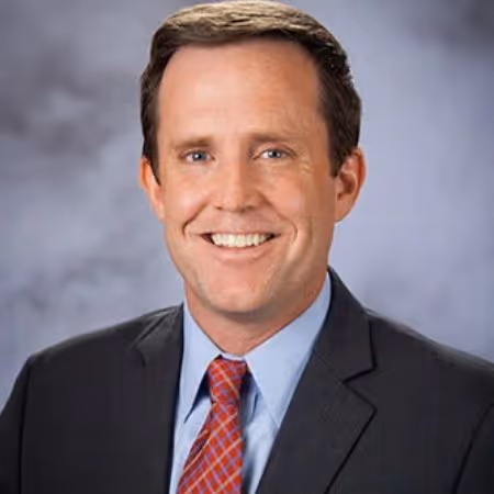 Smiling man wearing a dark suit, light blue shirt, and red patterned tie against a gray background.