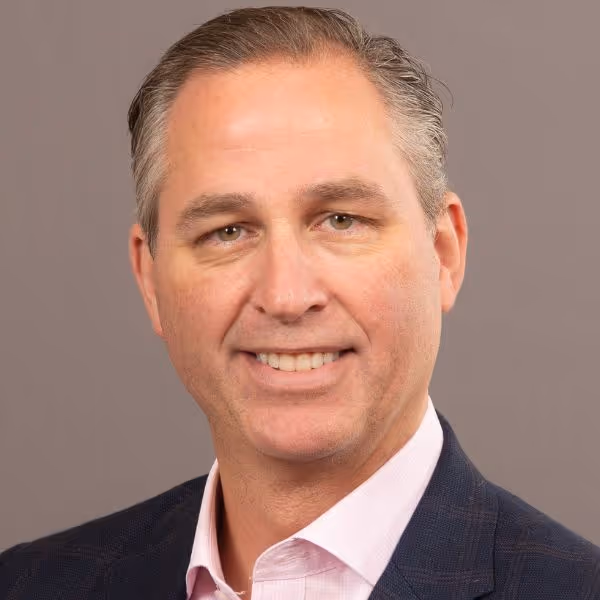 Smiling middle-aged man with short grayish hair wearing a dark blazer and light pink collared shirt against a plain background.