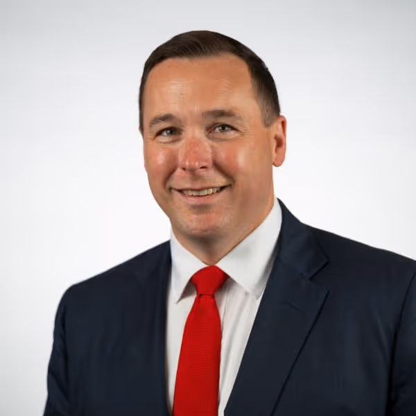 Man in a dark suit, white shirt, and red tie smiling against a plain white background.