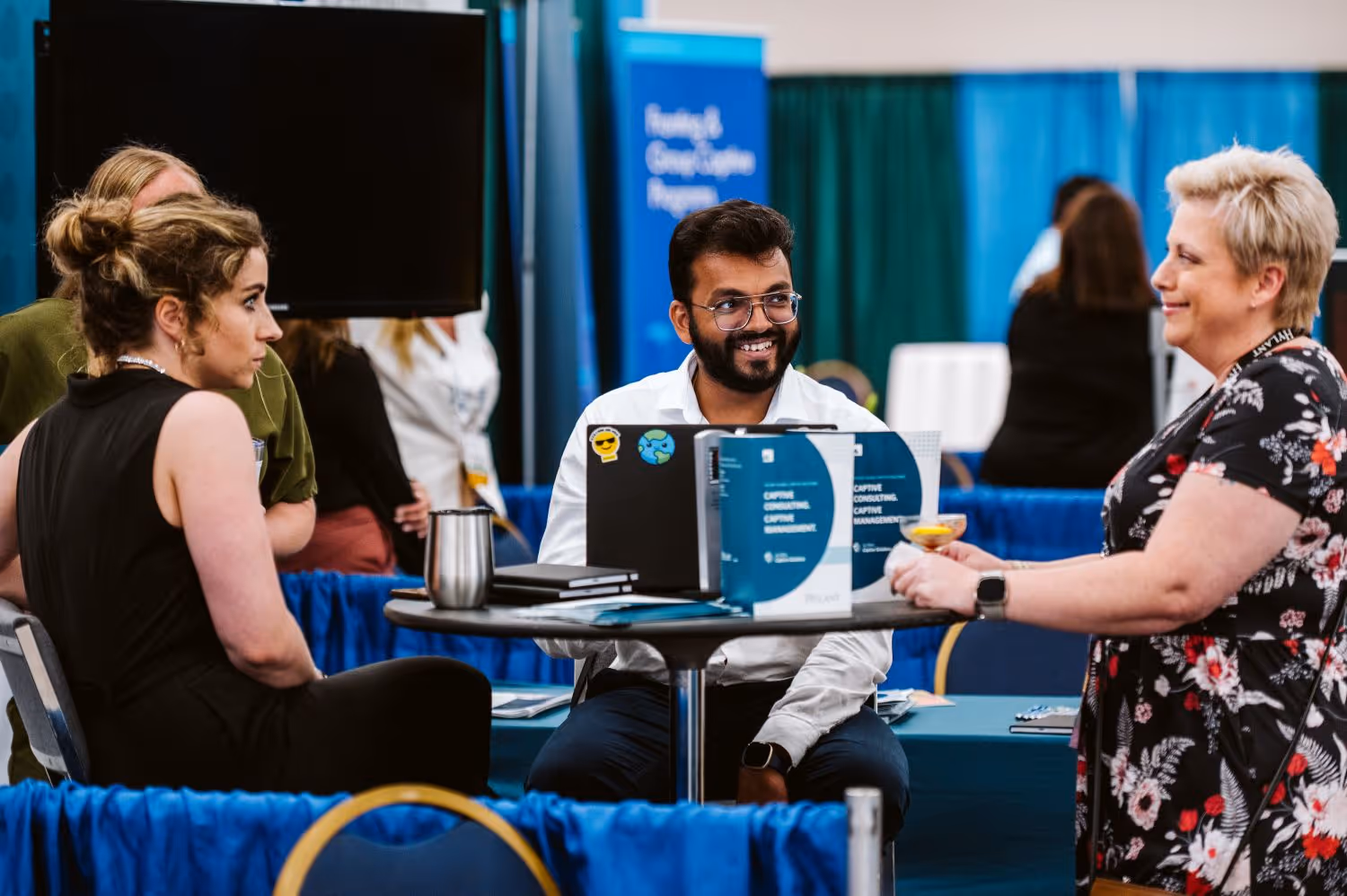 Three people engaged in a conversation at a round table during a professional event with informational brochures and laptops.