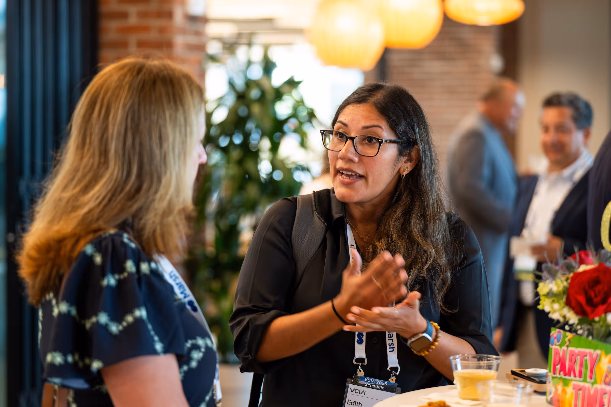 Two women engaged in a lively conversation at an indoor event with blurred people and decorations in the background.