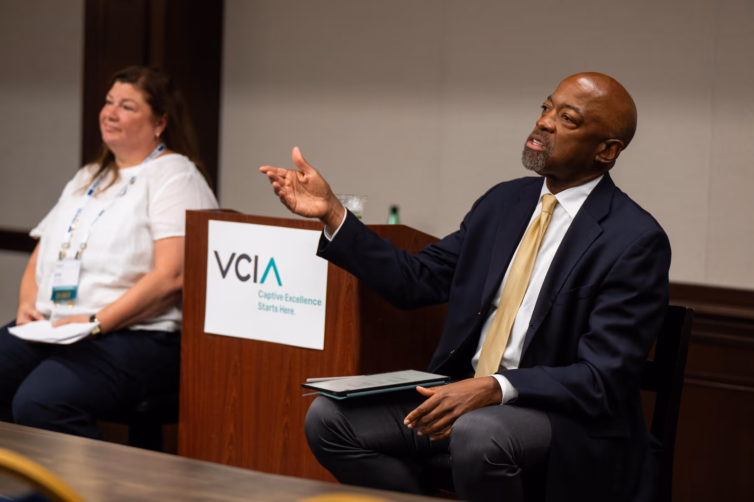Man in a suit and yellow tie speaking and gesturing with his hand during a panel discussion, with a woman seated beside him and a podium labeled VCIA in the background.