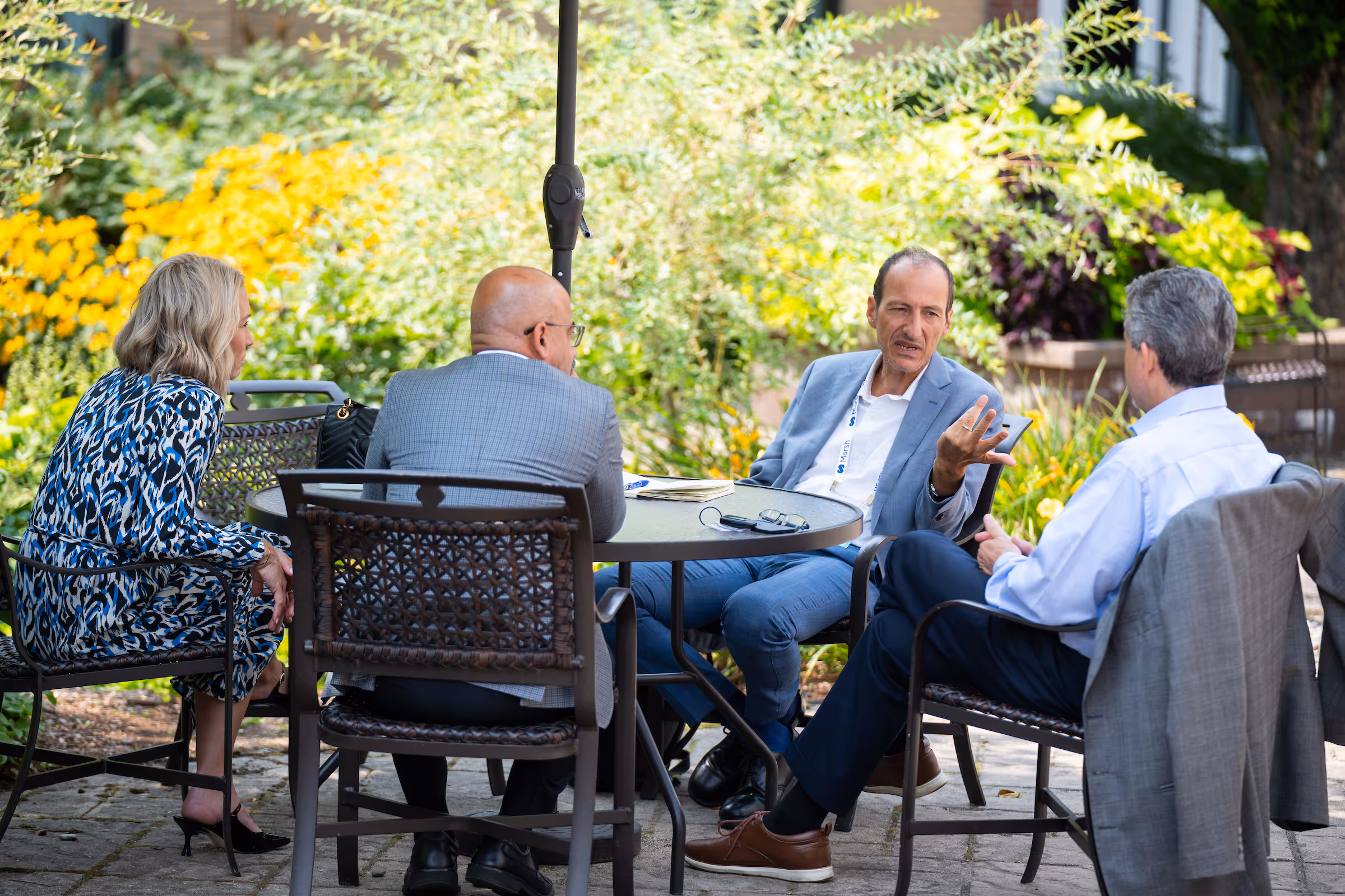 Four people in business attire seated around a round outdoor table engaged in conversation with greenery and yellow flowers in the background.