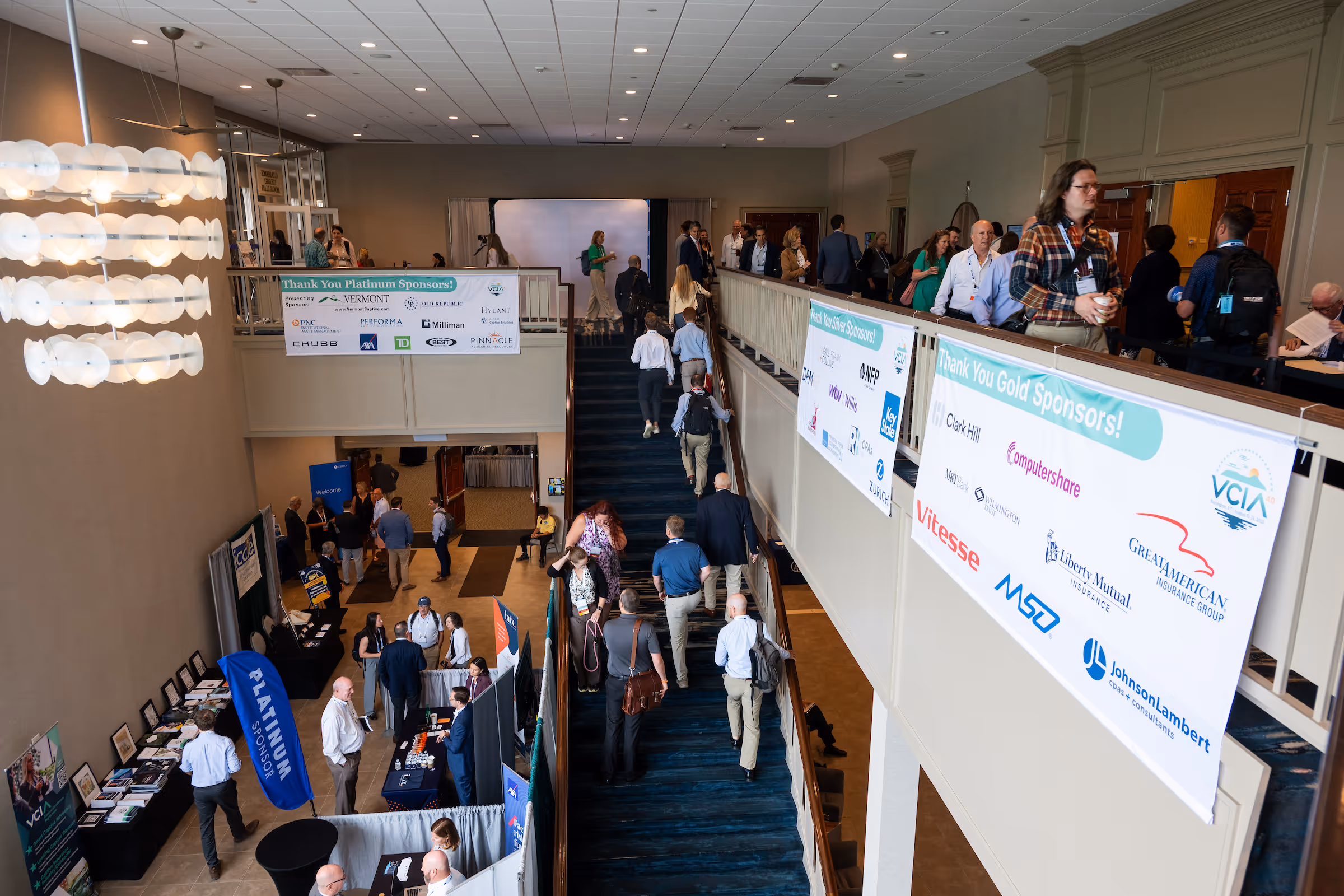 Conference attendees walking up stairs and mingling near sponsor booths and banners in a large venue.
