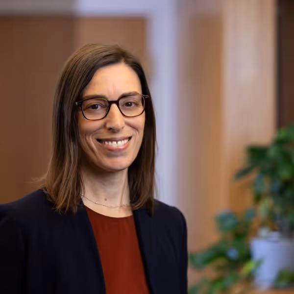 Smiling woman with straight brown hair, glasses, wearing a dark blazer and red top with an indoor blurred background.