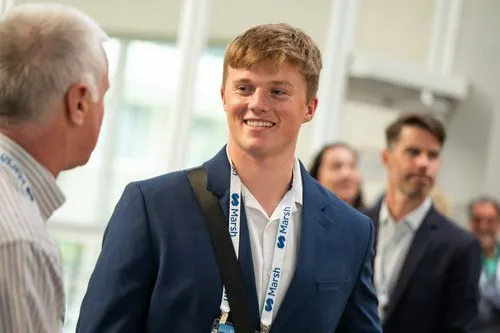 Young man in a blue suit and white shirt smiling and talking to an older man in a striped shirt at a professional event.
