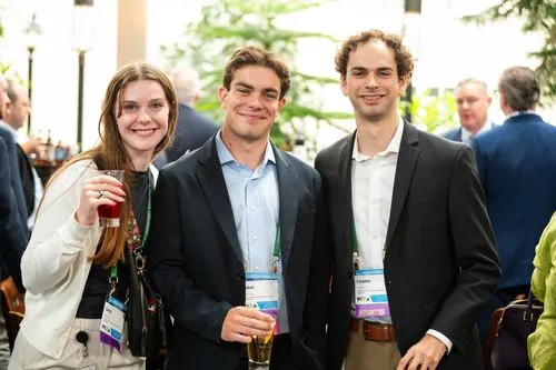 Three young adults smiling and posing with drinks at a professional event wearing name badges.
