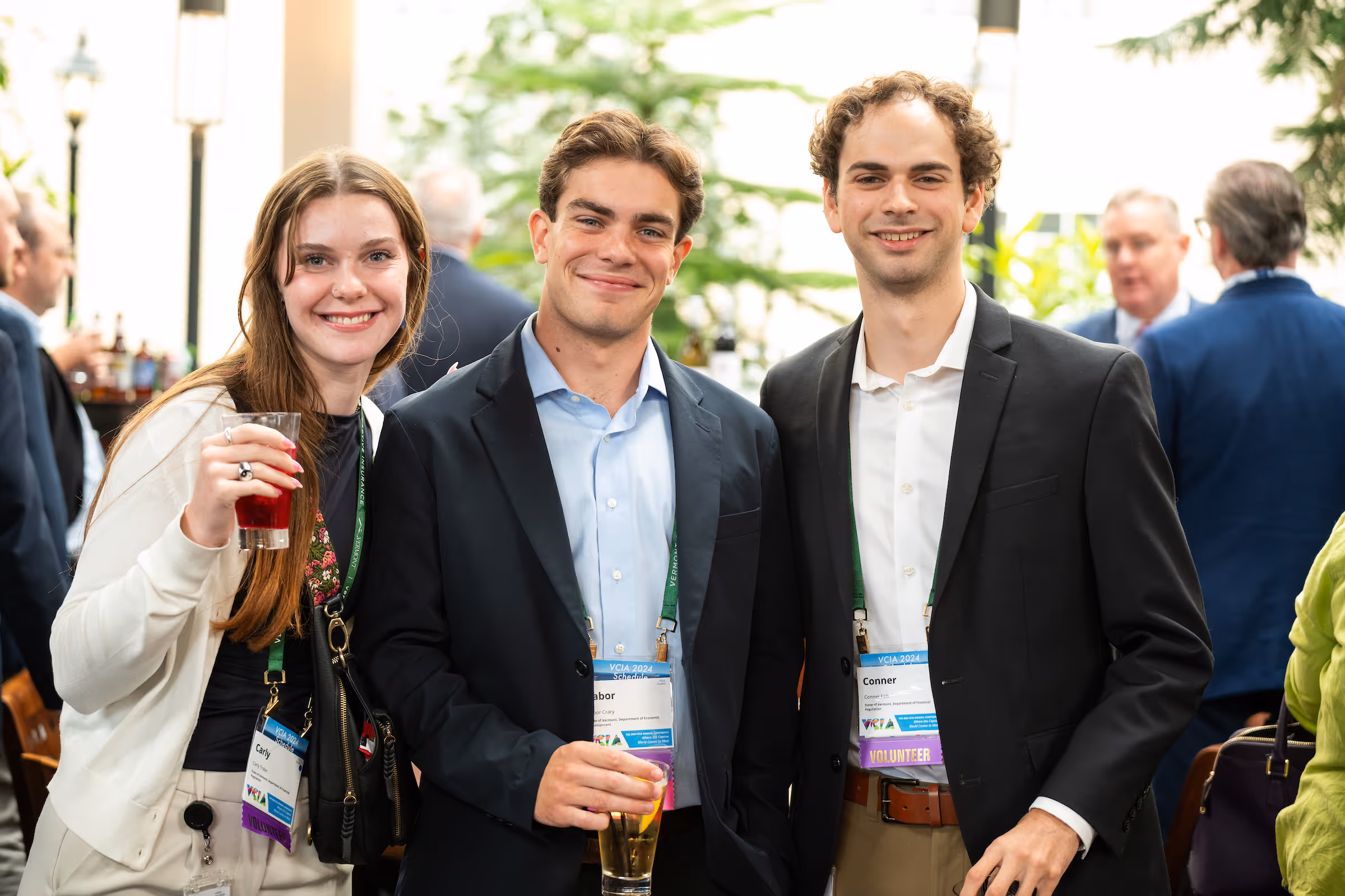 Three young adults smiling and posing with drinks at a professional event wearing name badges.