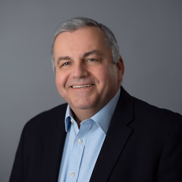 Smiling middle-aged man with gray hair wearing a light blue checkered shirt and dark blazer against a gray background.