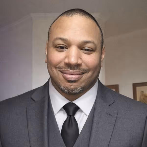 Smiling man wearing a dark gray suit, white shirt, and black tie against a neutral background.