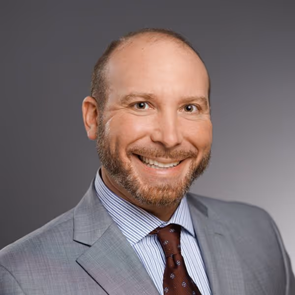 Smiling man in gray suit with striped shirt and patterned tie against a gray background.