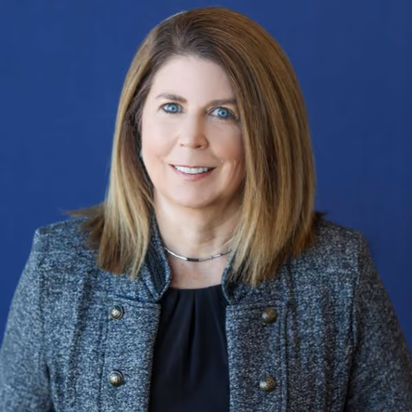 Smiling woman with shoulder-length light brown hair wearing a gray jacket and black top against blue background.
