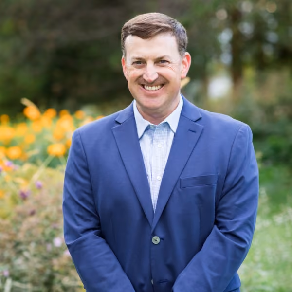 Smiling man in a blue suit jacket standing outdoors with blurred greenery and flowers in the background.