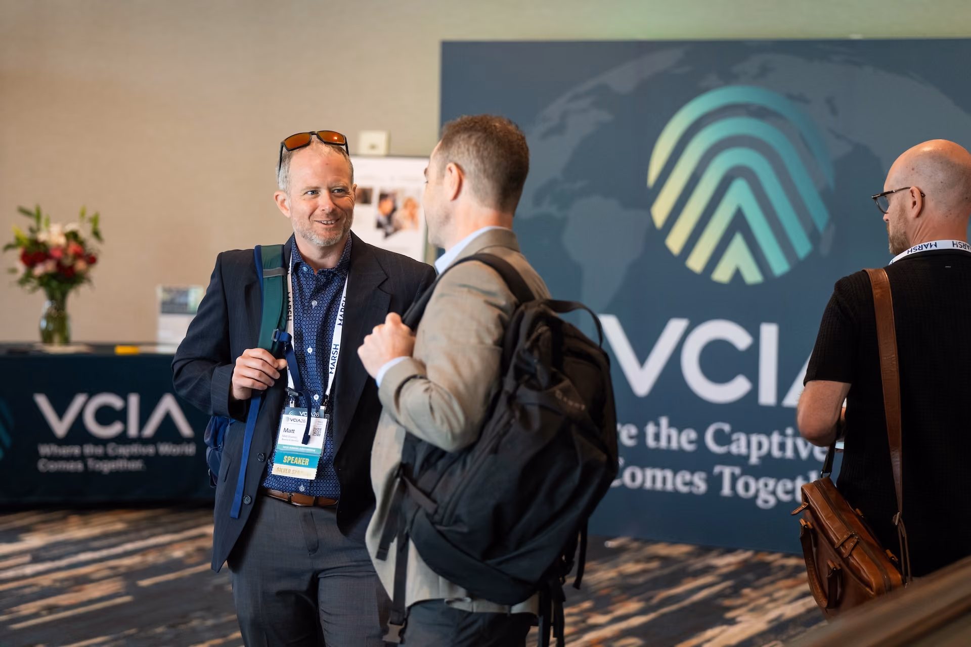 Three men talking indoors at a VCIA event, two with backpacks, one with speaker badge and sunglasses on head.