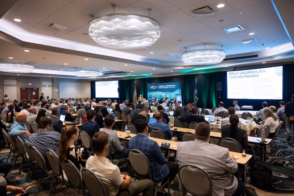 Conference room with a large audience seated at tables facing a panel of four speakers on stage beneath a banner reading 'VCIA Annual Conference'. Two projection screens display a presentation titled 'When Valuations Are Typically Needed'.