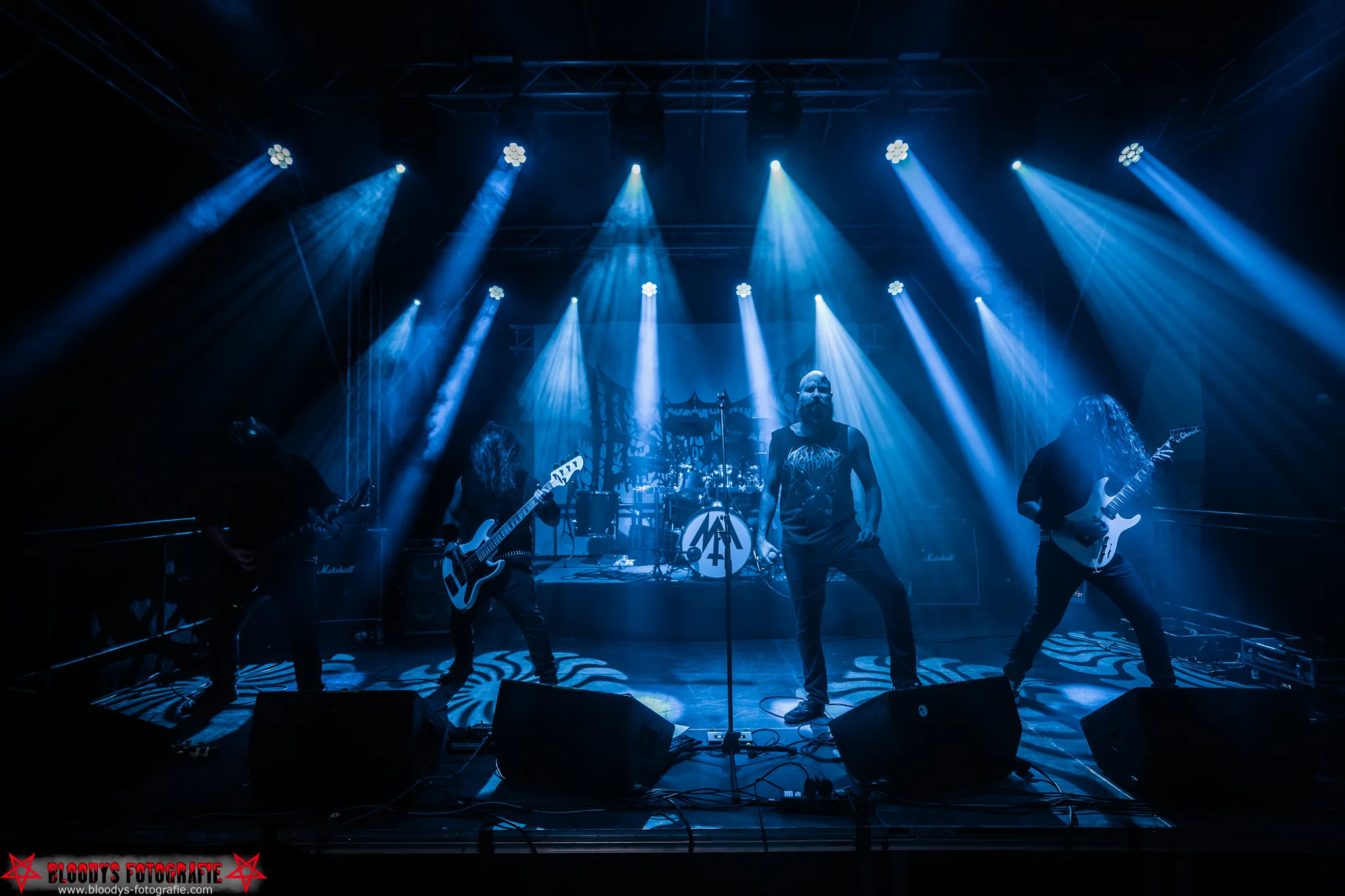 Metal band performing on stage under blue spotlights with guitars and drums.
