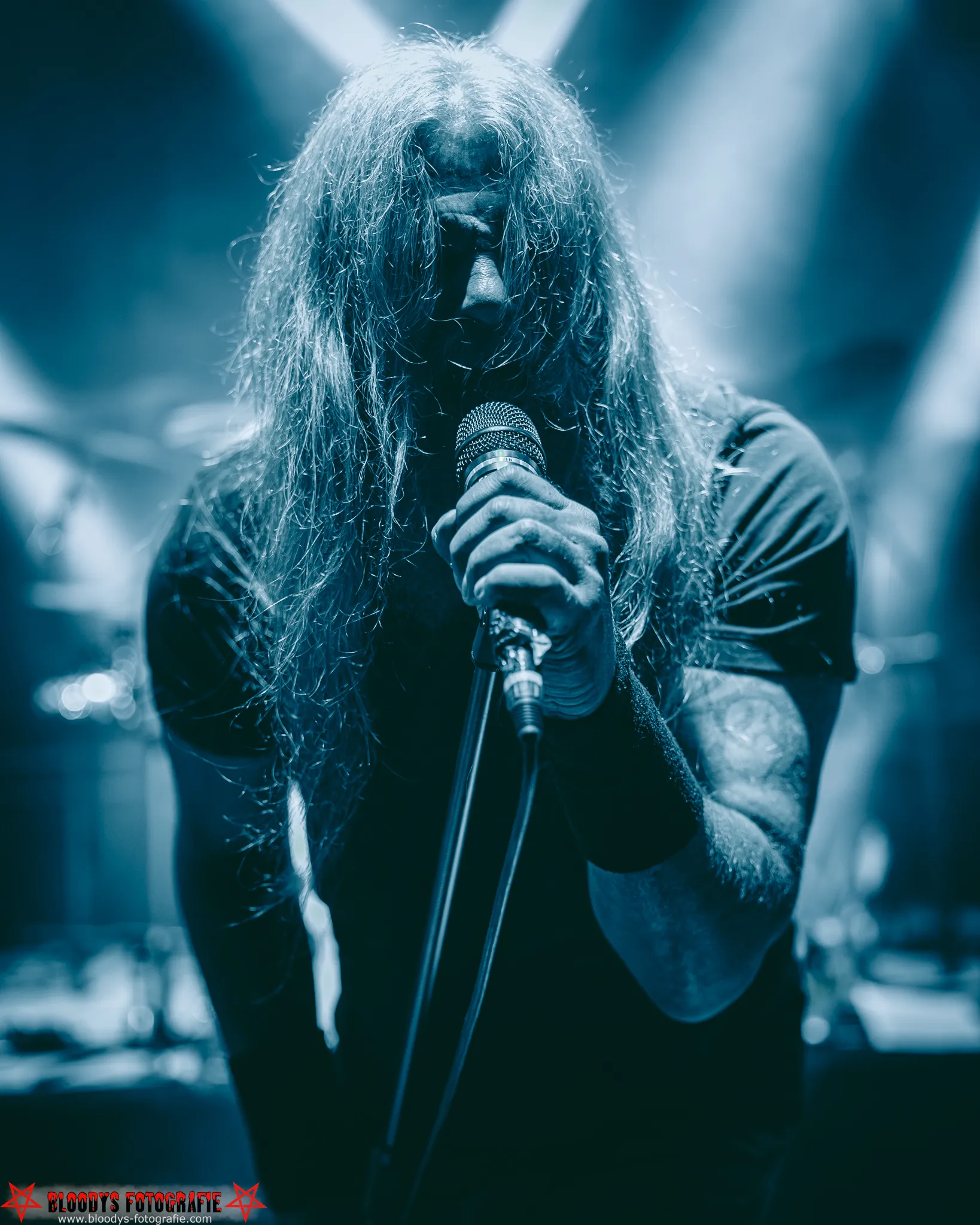 Long-haired singer gripping a microphone on stage with dramatic blue lighting beams in the background.